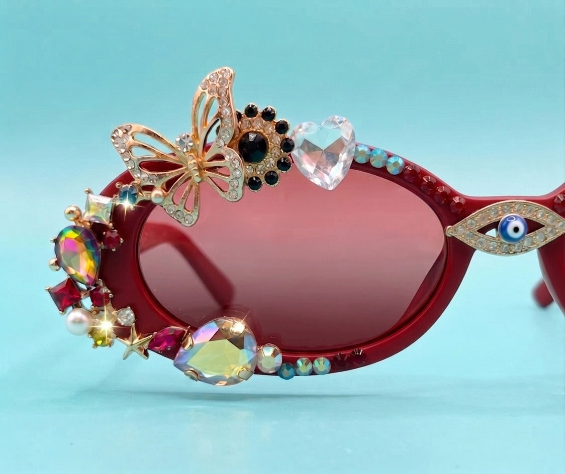 Red cat-eye sunglasses heavily embellished with decorative rhinestones, a gold butterfly ornament, a heart-shaped crystal, and an evil eye lens detail, set against a light blue background.