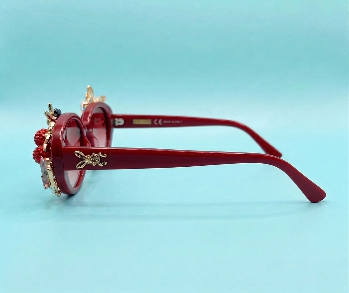 Red round-frame sunglasses with ornate gold-tone decorative details and red bead embellishments on the left side, shown on a light blue background.