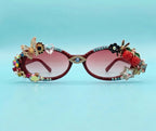Decorated cat-eye sunglasses with pink gradient lenses, featuring gold-tone metal frames embellished with rhinestones, a butterfly, a flower, a bee, and fruit charms including strawberries and a raspberry on a light blue background.