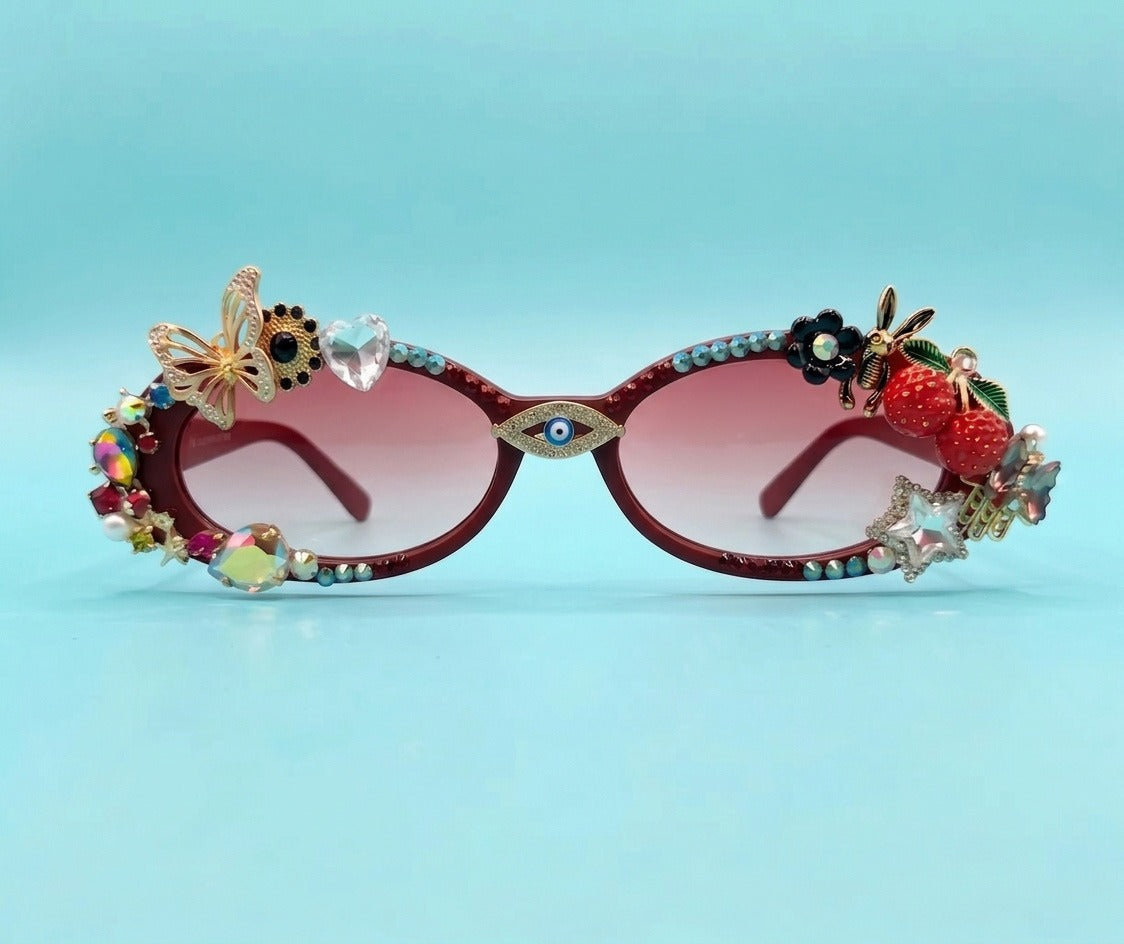 Decorated cat-eye sunglasses with pink gradient lenses, featuring gold-tone metal frames embellished with rhinestones, a butterfly, a flower, a bee, and fruit charms including strawberries and a raspberry on a light blue background.