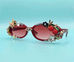 Decorated cat-eye sunglasses with pink gradient lenses, featuring gold-tone metal frames embellished with rhinestones, a butterfly, an eye motif, and 3D floral and fruit ornaments including a raspberry and star on a light blue background.